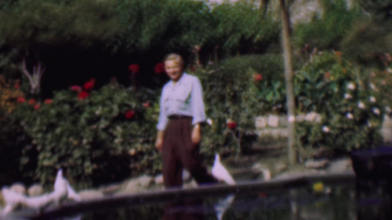 Smiling man stands near a pond with white pigeons and garden beds behind him.