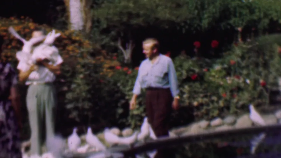A woman in light clothing holds white pigeons near the pond as a man walks nearby through the rose garden in the 1961 San Diego home-movie scene.