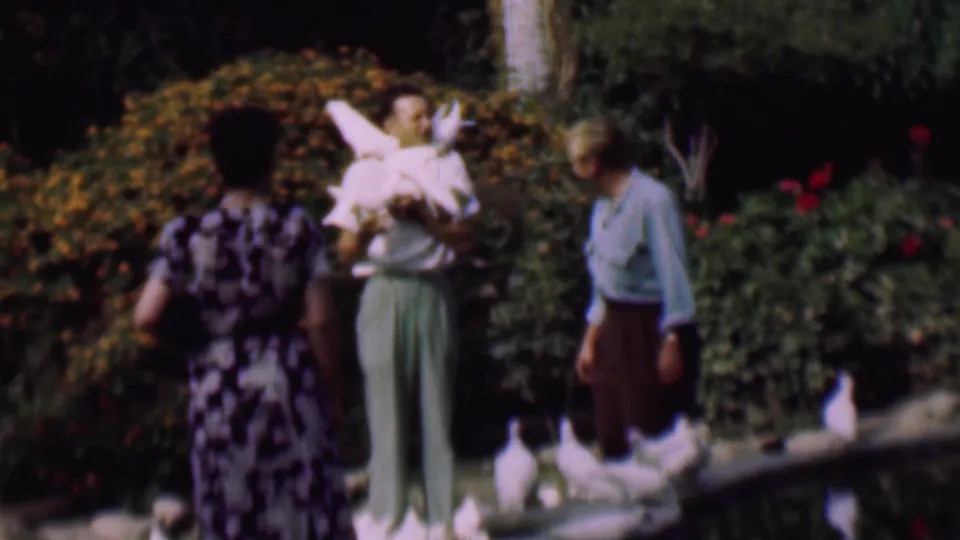Two adult visitors, including a woman holding white pigeons and a man watching nearby, stand by a pond in a 1961 San Diego garden scene.