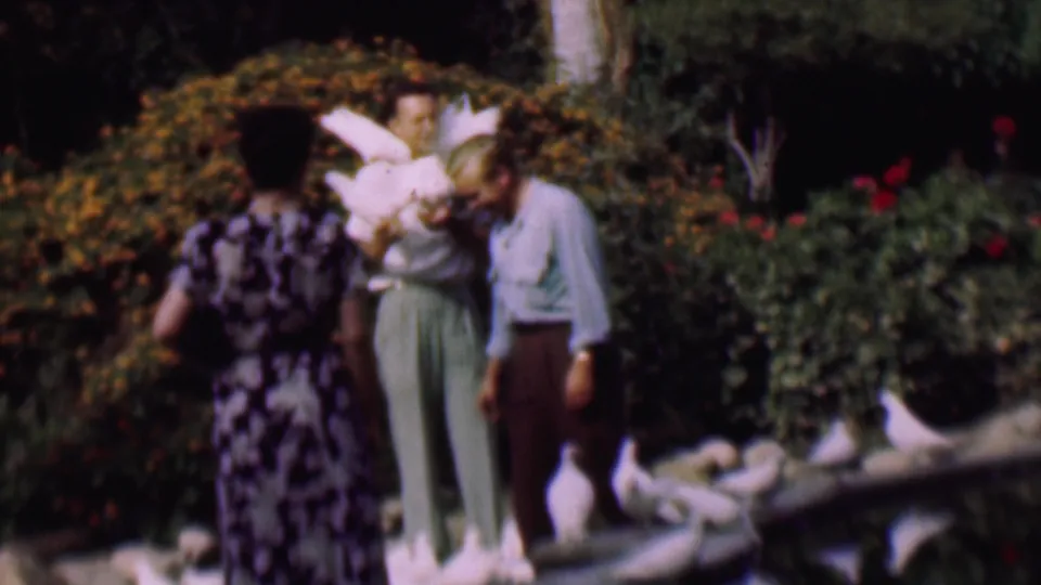 In a 1961 San Diego garden scene, a man holds white pigeons on his arms and shoulders while a woman in a patterned dress and another man stand nearby among shrubs and rocks.