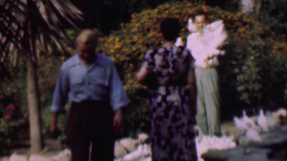 In the San Diego 1961 home-movie frame, a man in a blue shirt crosses the foreground while a woman and a man with white pigeons remain near shrubs and garden edging.