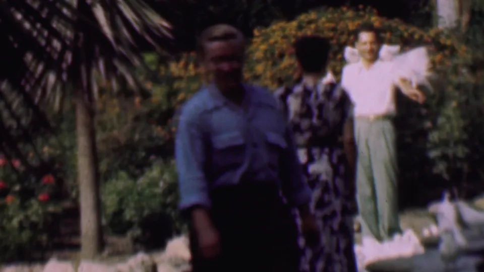 A 1961 San Diego garden frame shows a man in the foreground, a woman seen from behind, and a man in light trousers holding white pigeons near dense shrubs and rocks.