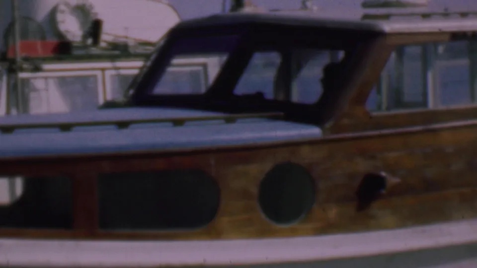 Close 1961 San Diego marina frame showing a classic wooden motorboat cabin, rounded porthole, pale deck, and a shadowed figure-like shape in the cockpit.