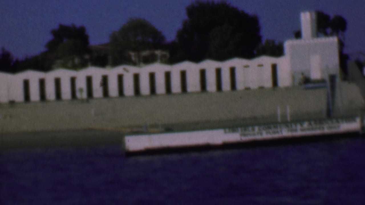 1961 San Diego home-movie frame showing a white stucco building across dark water, with arched openings, trees, lawn, and a small boat or platform marked with association text.