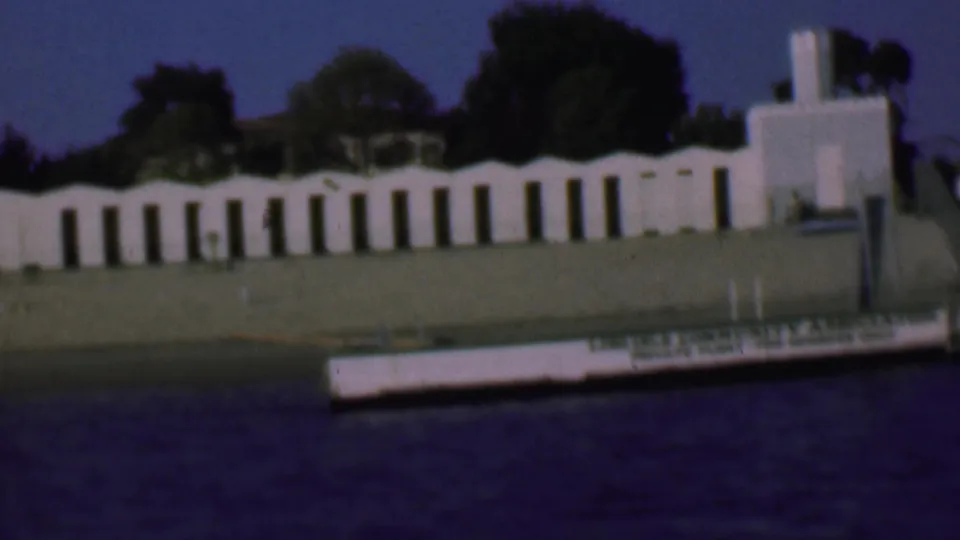 1961 San Diego home-movie frame showing a white stucco building across dark water, with arched openings, trees, lawn, and a small boat or platform marked with association text.