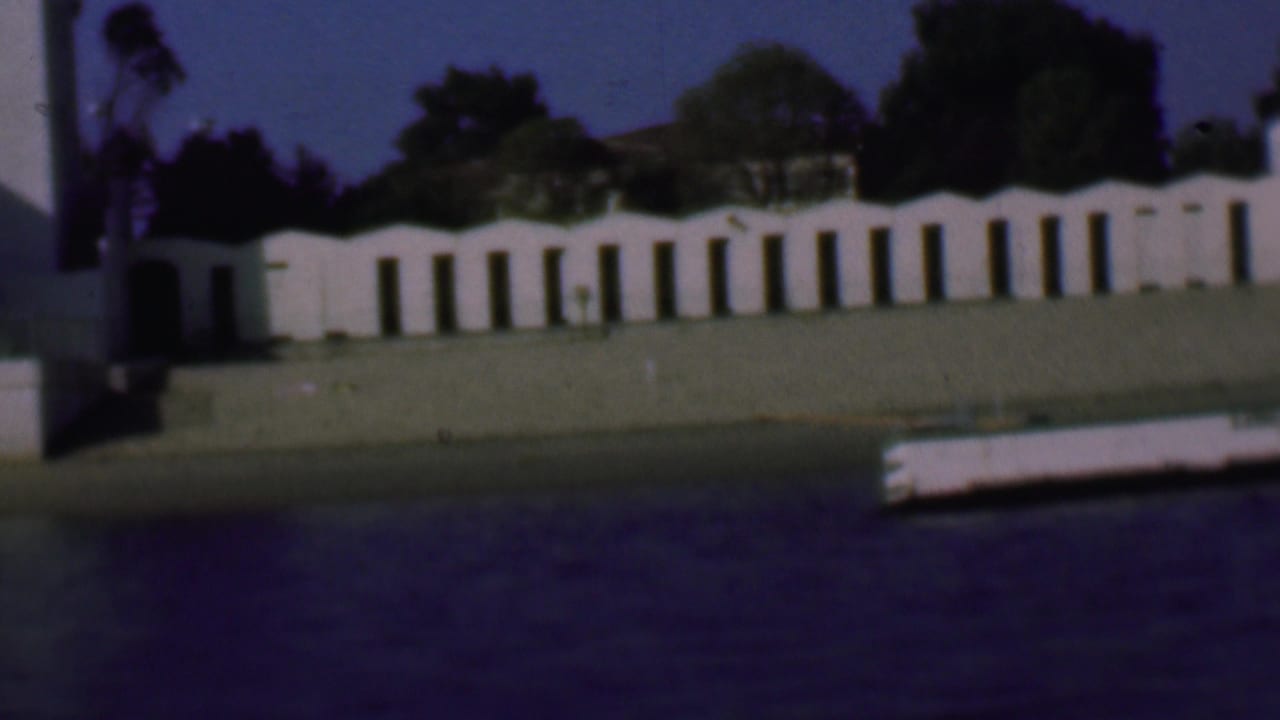 Long white wall with arched openings beside a grassy shoreline and water in the foreground.