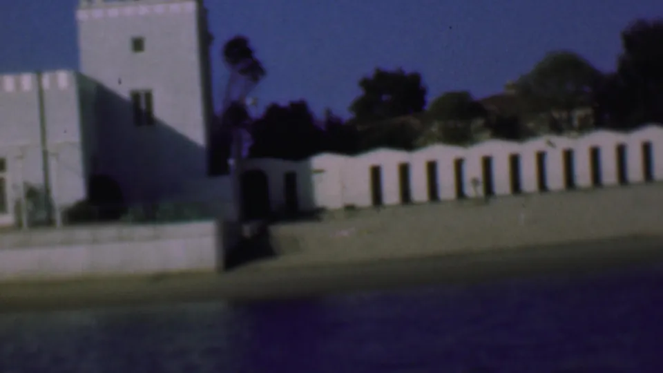 1961 San Diego home-movie frame with a white crenellated tower, arched stucco wall, shadowed entrance, trees, lawn, and waterfront embankment.