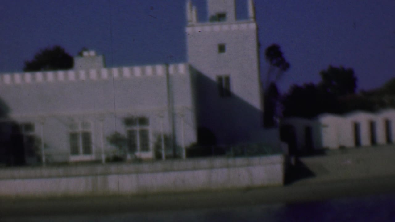 1961 San Diego home-movie frame showing a white stucco building with crenellated roofline, tower-like section, windows, low wall, and dark trees in the background.