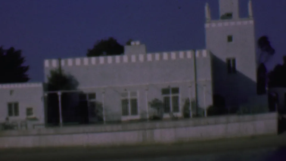 1961 San Diego frame of the same white stucco architectural complex, with a crenellated parapet, tower, foreground wall, shaded doorway area, and trees against a blue sky.