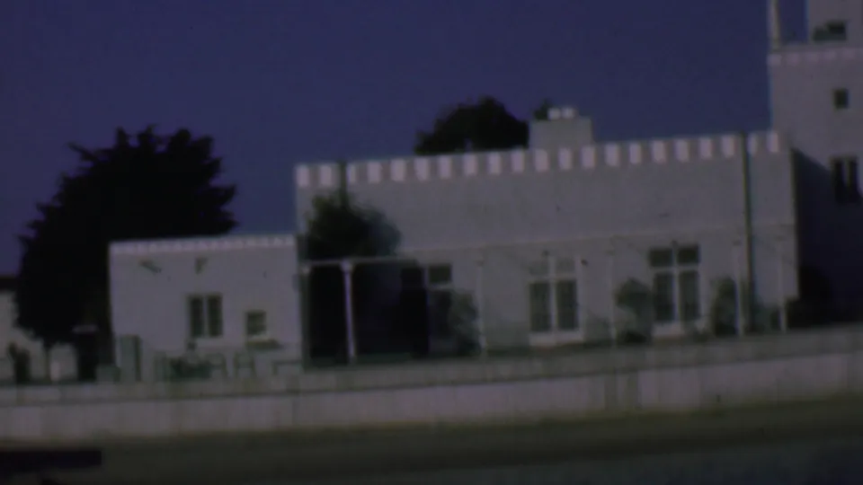1961 San Diego home-movie frame showing a panned view of the white stucco building, with the tower partly at the right edge, a smaller adjoining section, windows, trees, and low wall.