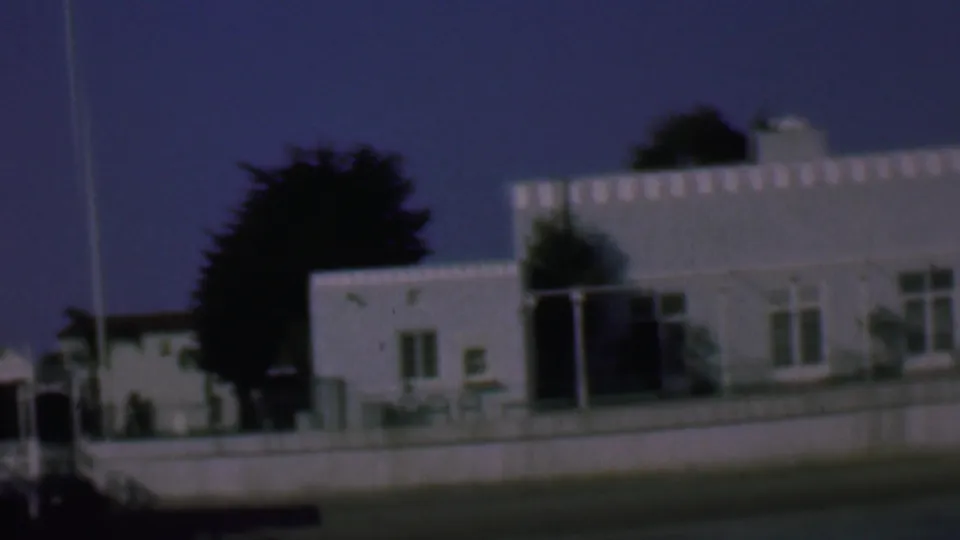 Final 1961 San Diego frame showing the white stucco building partially cropped, with a vertical pole or mast in front, low wall, dark tree shapes, and deep blue sky.
