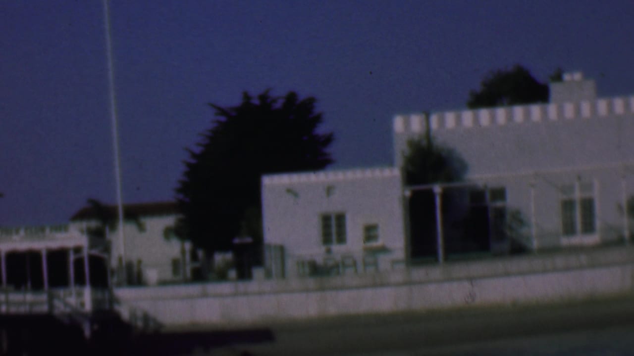 1961 San Diego home-movie frame showing a white waterfront building complex with railings, a dark tree behind it, and a deep blue sky.