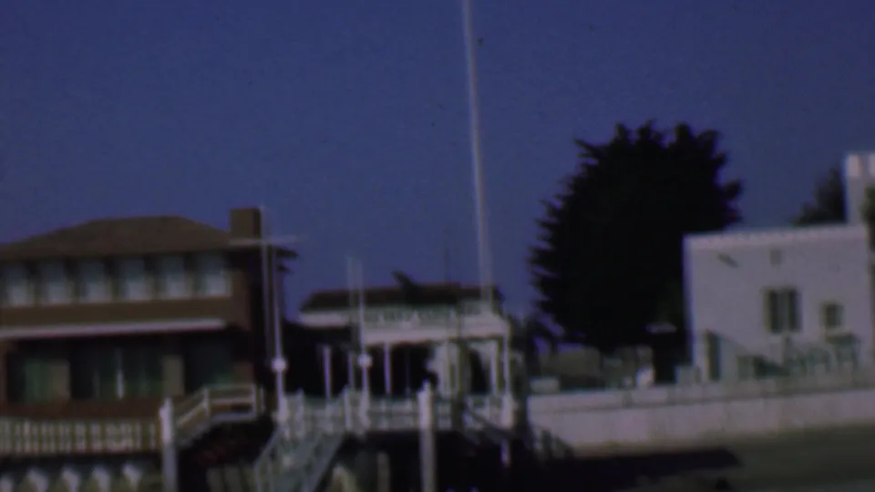 1961 San Diego frame showing a brown waterfront building next to white structures, railings, a flagpole, and a dark tree in the background.