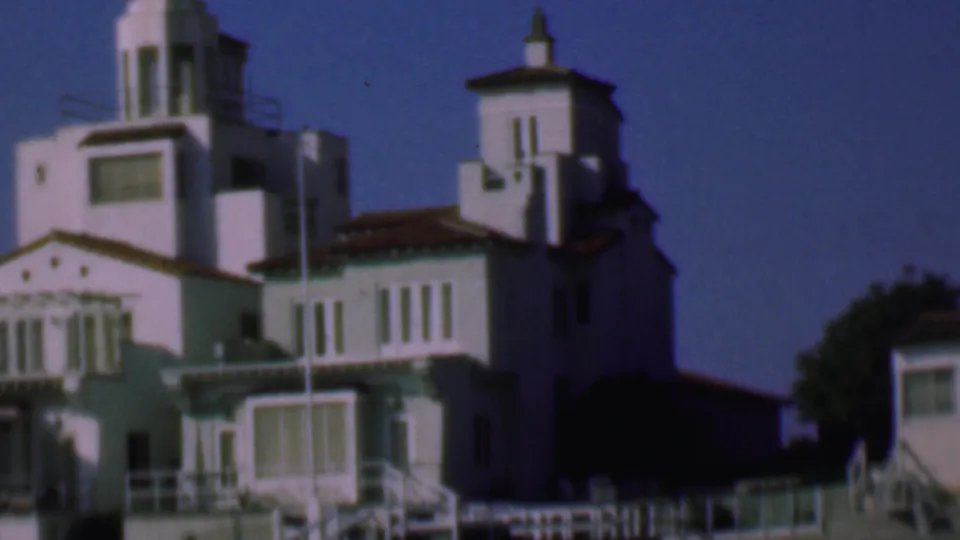 1961 San Diego home-movie frame showing a larger white building with tower-like sections, red-tile roof details, railings, and a deep blue sky.