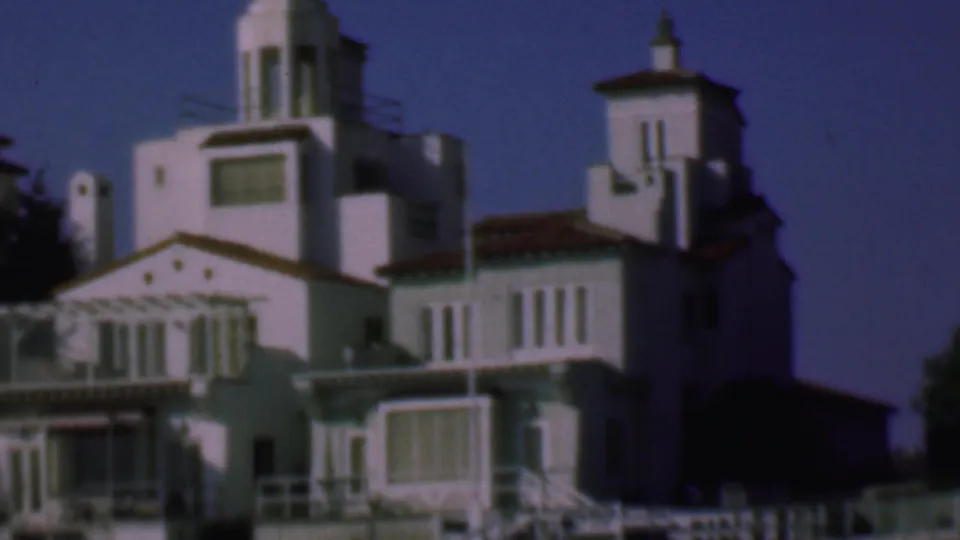 1961 San Diego home-movie frame showing white stucco residential buildings with red tile roofs, balconies, shuttered windows, and tower forms under a deep blue sky.