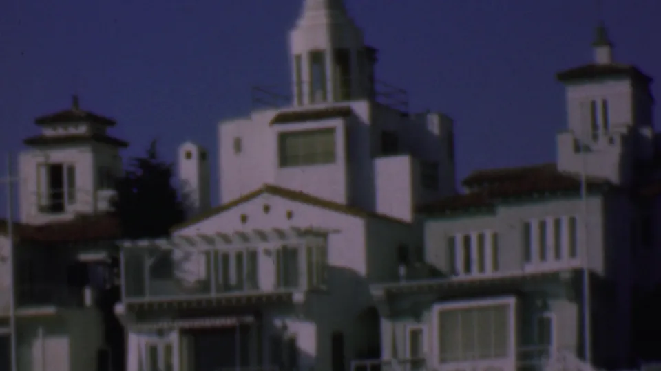 1961 San Diego home-movie still showing a row of white stucco houses with roof towers, narrow windows, balcony railings, and shaded lower facades.