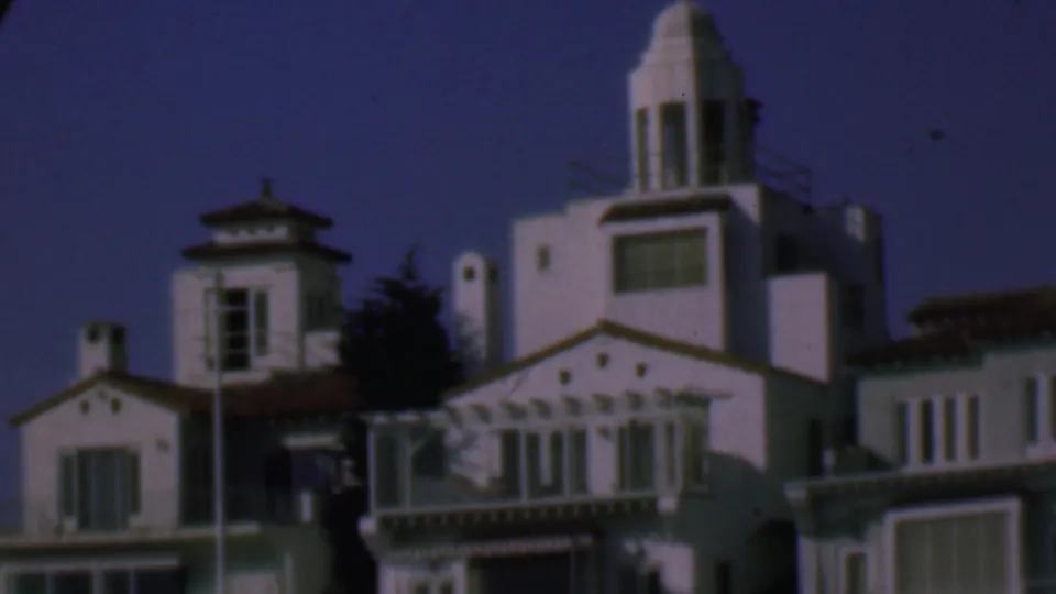 1961 San Diego frame showing the pan continuing across white Spanish-style residential exteriors, with towered rooflines, shuttered windows, red tile accents, and trees in shadow.