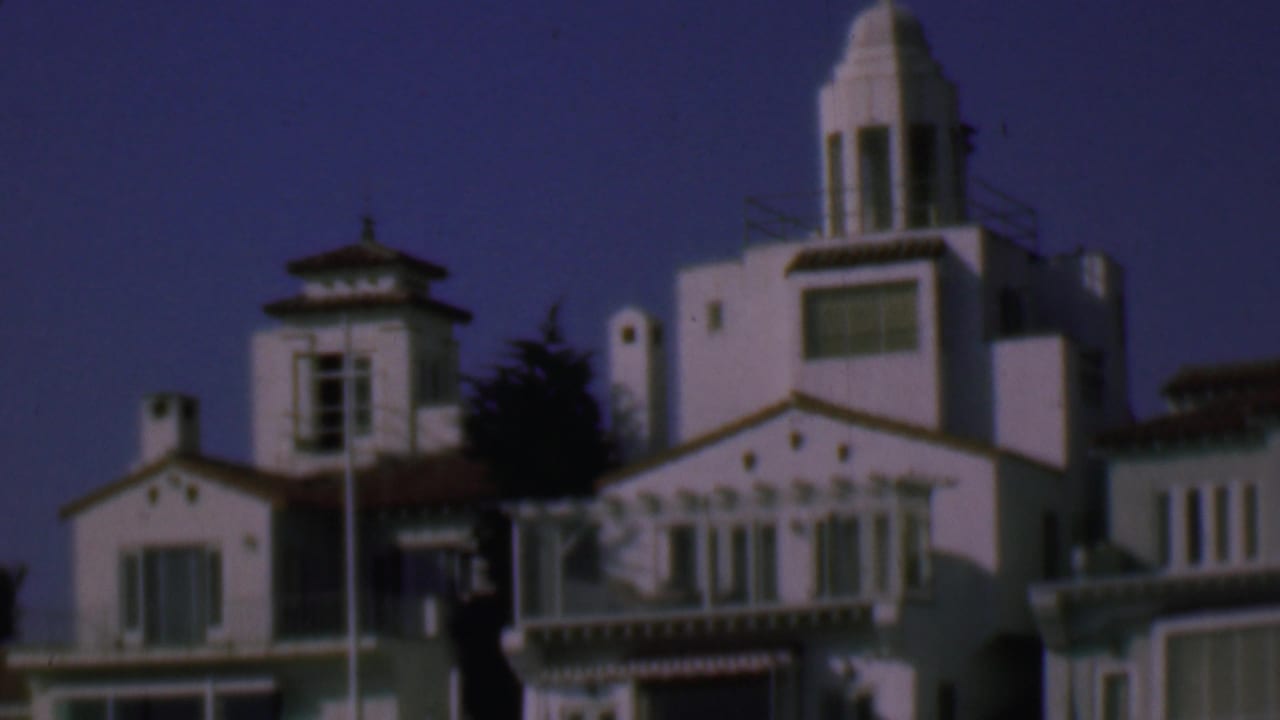1961 San Diego home-movie frame showing two whitewashed Spanish-style multistory buildings with red tile roof edges, balconies, towers, trees, and deep blue sky.