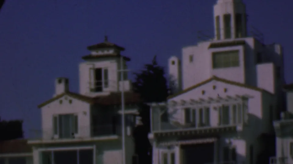 1961 San Diego frame with the camera centered on white stucco Spanish-style facades, ornate balcony trim, tall tower elements, windows, and dark trees in the background.