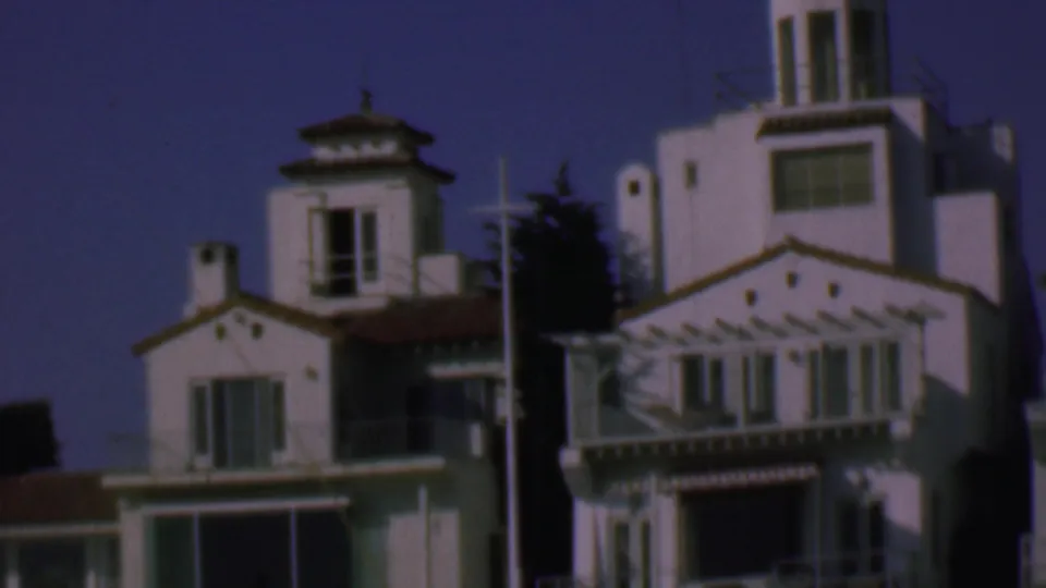 1961 San Diego home-movie frame showing a closer view of Spanish-style stucco architecture with a tall tower, red tile roofline, balcony details, windows, and a vertical pole.