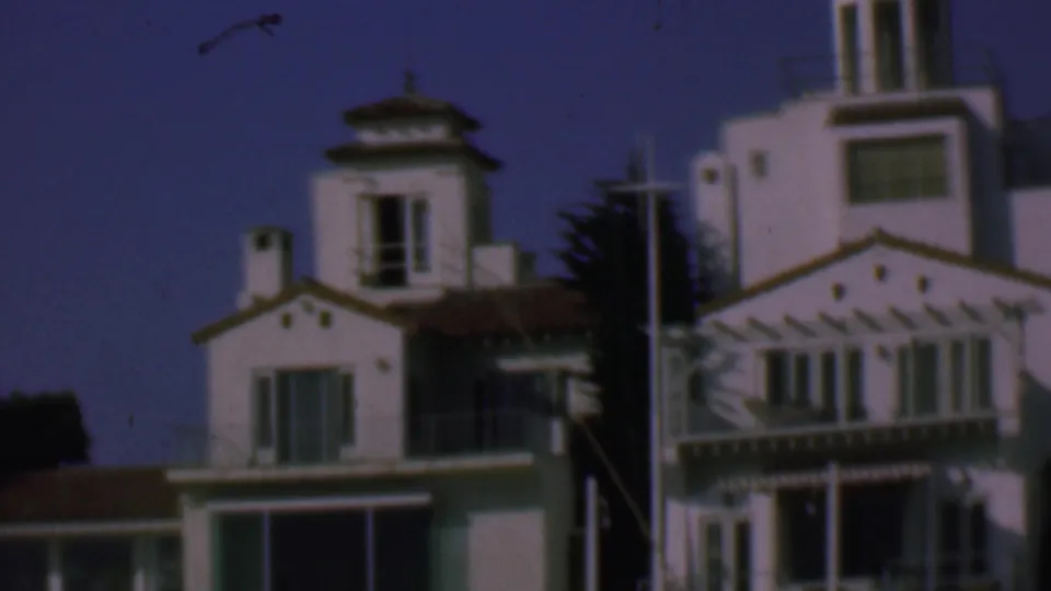 1961 San Diego frame showing whitewashed Spanish-style buildings with terracotta rooflines, balcony structures, dark trees, window openings, and a pale pole in the foreground.