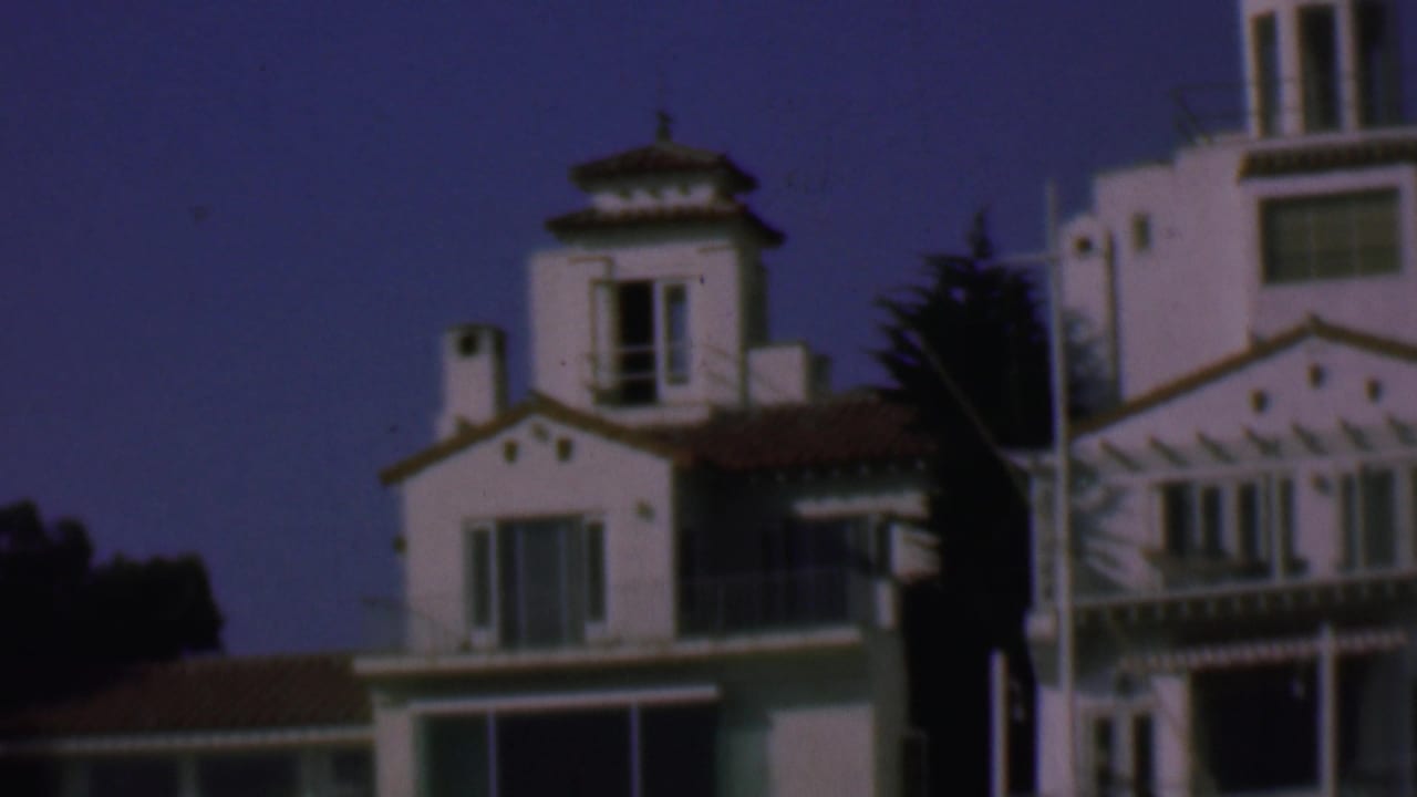 1961 San Diego home-movie frame showing white Spanish-style waterfront houses with terracotta rooflines, balconies, windows, and dark trees behind them.