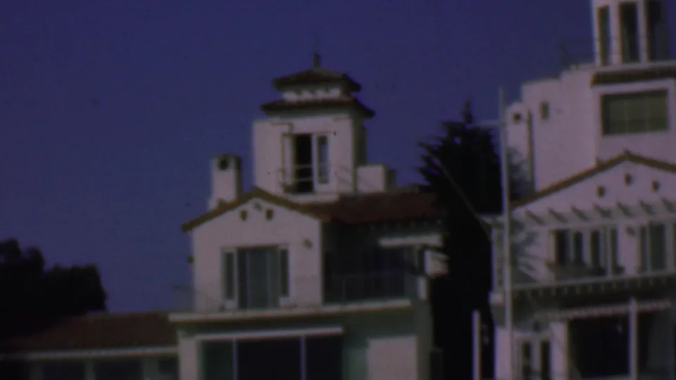 1961 San Diego home-movie frame showing white Spanish-style waterfront houses with terracotta rooflines, balconies, windows, and dark trees behind them.