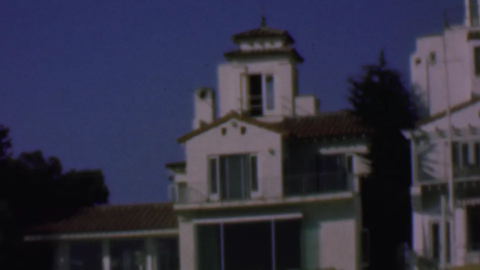 1961 San Diego frame with white stucco beach houses, small balconies, shuttered windows, tiled roof edges, and trees visible in the background.