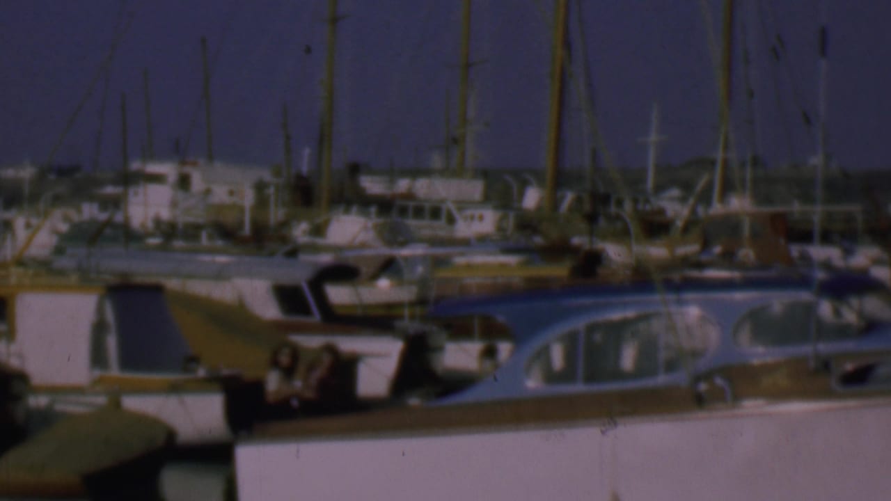 1961 San Diego home-movie frame showing a marina with white cabin boats, tall sailboat masts, dock structures, and a few small indistinct people near the boats.