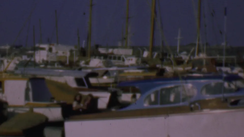 1961 San Diego home-movie frame showing a marina with white cabin boats, tall sailboat masts, dock structures, and a few small indistinct people near the boats.