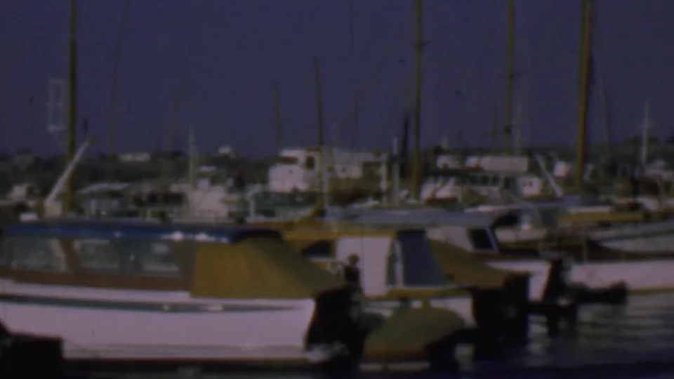 1961 San Diego home-movie frame centered on a white cabin boat with a yellow cover, with marina water, masts, other boats, and distant dockside buildings behind it.