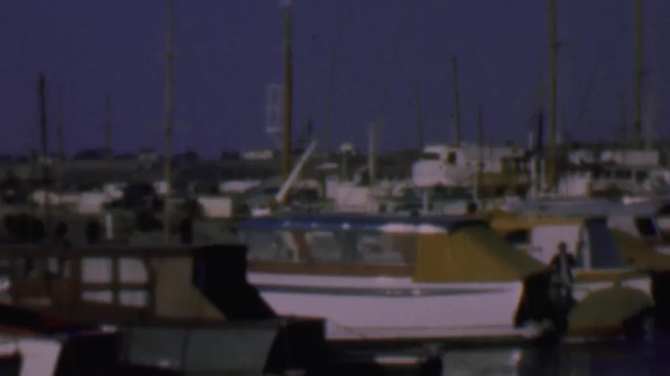 1961 San Diego marina frame showing docked cabin boats, vertical sailboat masts, low background buildings, calm water, and a small indistinct person near a boat at the right.