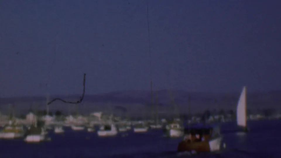 Wider San Diego bay view from the 1961 8mm clip, showing open water, distant moored boats, clustered masts, shoreline haze, and a white sail at the right edge.