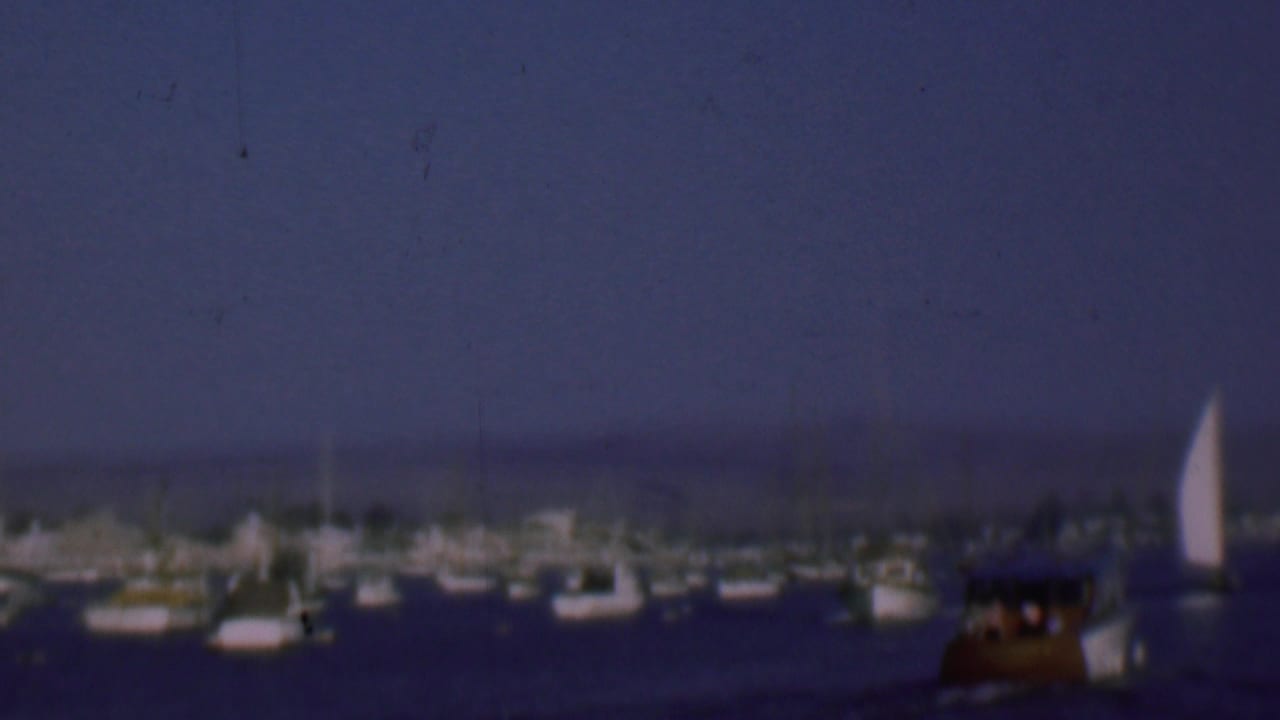 1961 San Diego home-movie frame showing rows of anchored sailboats and small motorboats on dark harbor water beneath a blue dusk sky.