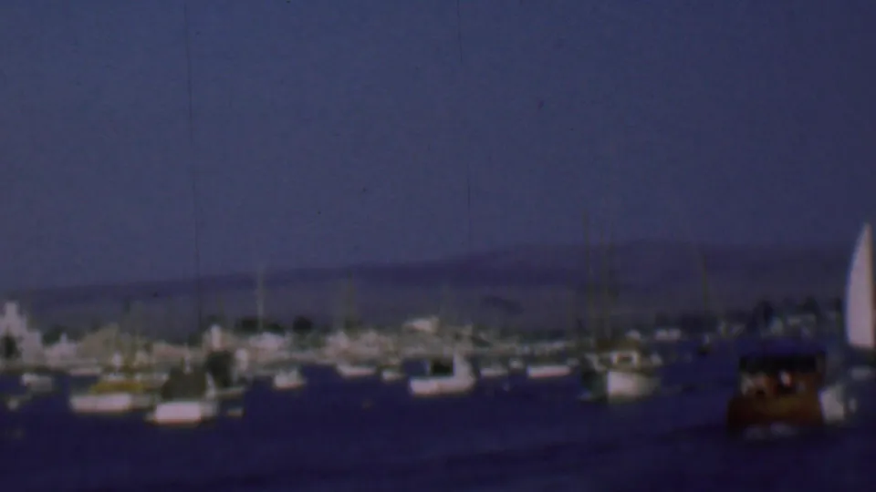 Soft 8mm frame of the San Diego marina in 1961, with clustered white sailboats, calm water, and a low horizon of hills and shoreline structures.