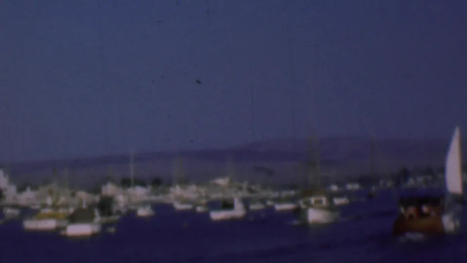 1961 San Diego harbor frame showing a bright white sail at the right edge, rows of anchored boats, dark water, and distant hills in a grainy dusk view.
