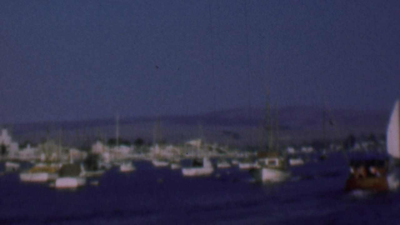 1961 8mm San Diego harbor view with sailboats and small vessels anchored in calm water beneath a deep blue sky.