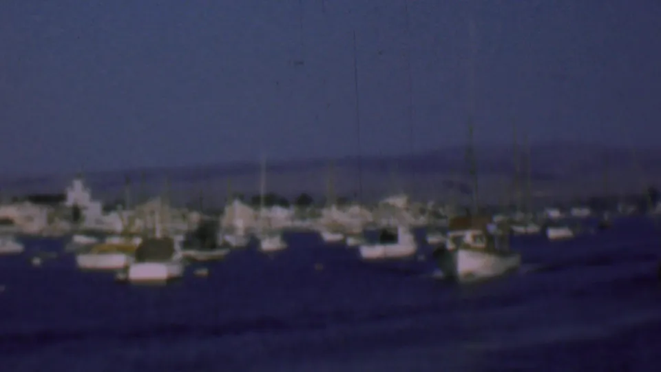 Home-movie frame from 1961 San Diego showing sailboats and small vessels spread across calm bay water with a faint shoreline beyond.