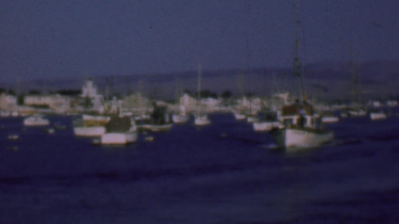 1961 San Diego harbor view with a small fishing boat in the foreground, other boats scattered across calm water, and a hazy shoreline beyond.