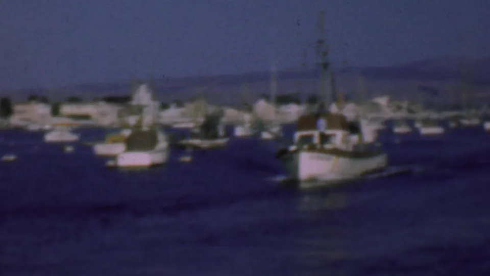 A small fishing boat passes through San Diego harbor water, with clustered vessels, a faint wake, and hazy hills visible in the background.