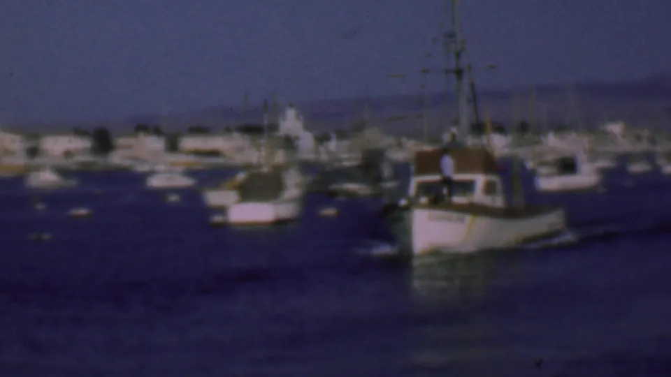 Fishing boat with small crew figures visible on deck heads through San Diego harbor in 1961, with other boats and the waterfront behind it.