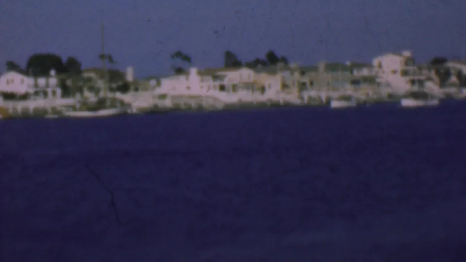 1961 San Diego waterfront with low buildings, small boats near shore, masts, trees, and dark harbor water captured in soft 8mm focus.