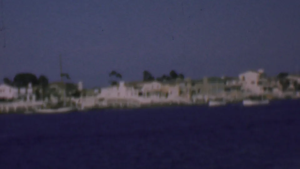 Soft 8mm frame of the San Diego harbor shoreline in 1961, showing moored boats, low coastal buildings, masts, and open water.