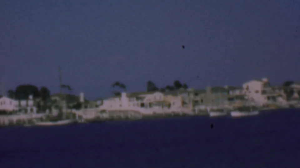 1961 San Diego harbor shoreline seen from across the water, with small boats, pale waterfront buildings, trees, and vertical masts softened by home-movie focus.