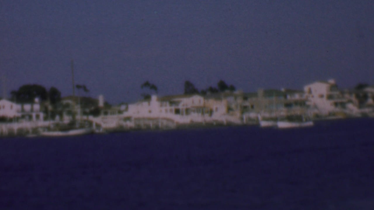 1961 San Diego-area waterfront view with pale shoreline homes, moored small boats, and dark blue water in soft 8mm color.