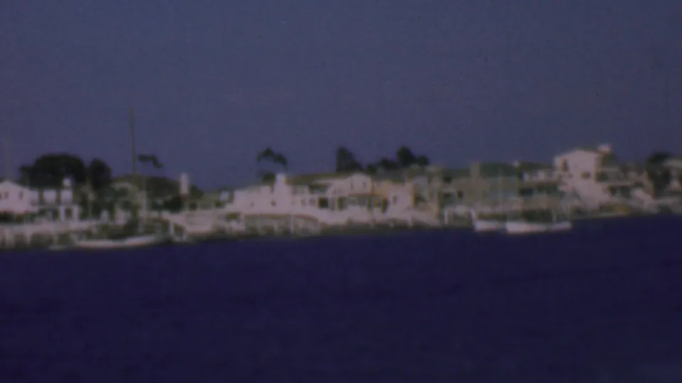 1961 San Diego-area waterfront view with pale shoreline homes, moored small boats, and dark blue water in soft 8mm color.