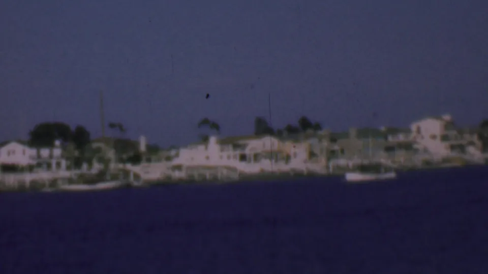 Distant San Diego waterfront in 1961, showing small moored boats, pale houses, docks, and a soft blue 8mm sky.