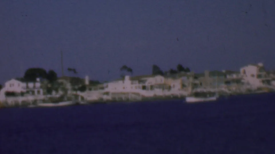 Soft 8mm frame of the supplied 1961 San Diego waterfront, with residential buildings, docked boats, and dark rippled water.