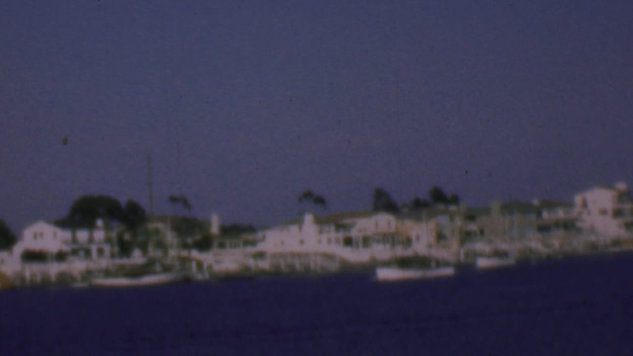 1961 San Diego coastal waterfront view from a moving boat, showing pale houses, moored small boats, trees, and open sky across the water.