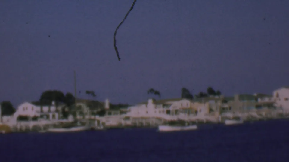 Soft 8mm frame of the San Diego shoreline in 1961, with white waterfront houses, a few small boats, trees, and aged-film scratches visible.